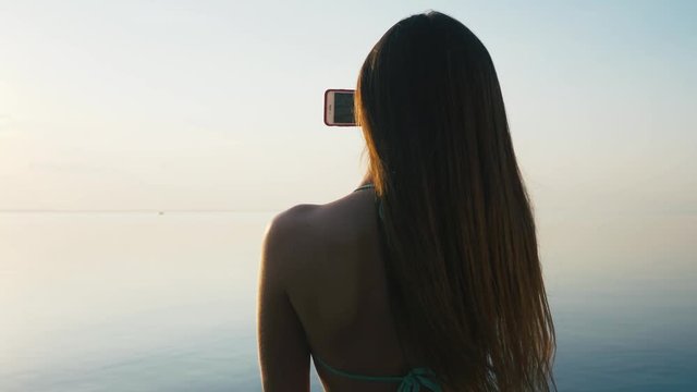 Pretty Young Woman Taking Pictures with her Smartphone at Sunset on the Beach near the Sea on Vacation. Slow Motion