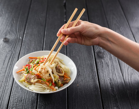 Slender woman hand with chopsticks reaching for Asian udon noodles with meat and vegetables on black background