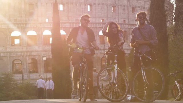 Three young friends tourists with bikes in colle oppio park in front of colosseum on road with trees at sunset having fun talking laughing in Rome slow motion lens flare
