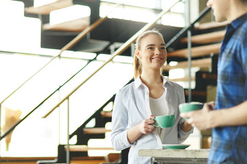 Pretty young woman and crop man standing in cafeteria with coffee cup and chatting happily.