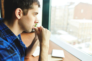 Side view of young man sitting at table with notepads and looking away through window contemplating on issue.