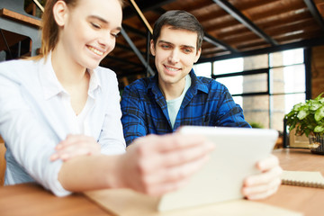 Cheerful content girl and boy sharing tablet sitting at table with studies and spending time together.