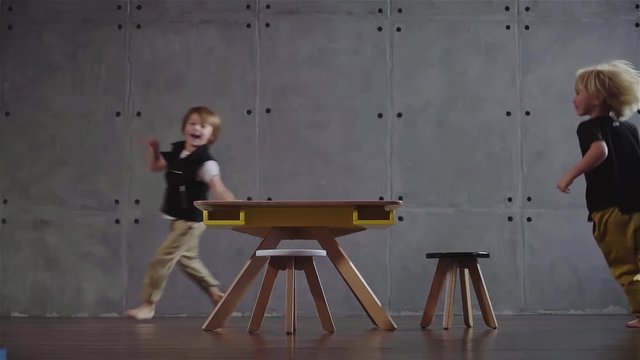 Two little boys with blond and dark hair running around a small kids table and laughing. A room with gray walls. Concept of happiness and carefree childhood time. Locked down slow motion medium shot