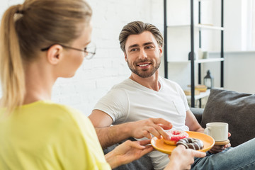 woman in eyeglasses holding plate with donuts while her boyfriend taking one donut and sitting on sofa at home