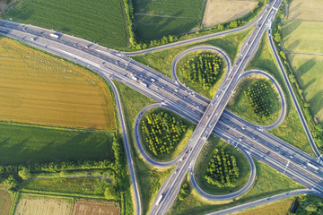 Cloverleaf interchange seen from above. Aerial view of highway road junction in the countryside with trees and cultivated fields. Bird's eye view. - obrazy, fototapety, plakaty