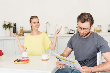 young woman with wide arms looking at boyfriend while he reading newspaper at table with breakfast