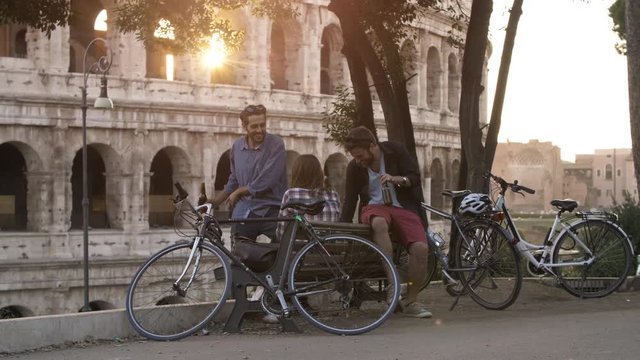 Three young friends tourists with bikes sitting on bench in front of colosseum under tree at sunset drinking beers having fun talking laughing chilling in Rome slow motion