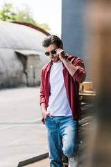 front view of young stylish man standing near wooden pallets and adjusting sunglasses