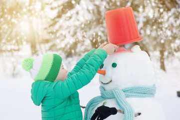 Little boy puts a bucket on the snowman's head