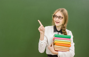 Smiling girl  with books standing near school board and pointing away. Space for text