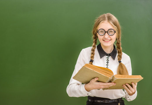 Smiling nerd student girl  with open book standing near green school board. Space for text