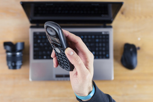Hand Holding a Cordless Telephone with Laptop, Mouse and Camera Lens in Background