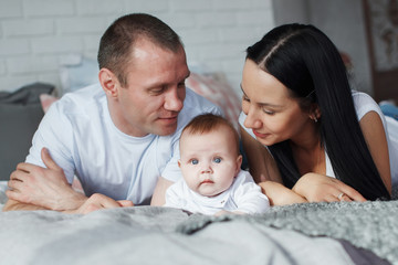 Fototapeta premium Young mom and dad and their baby son lie in bed and look at the camera indoors. The concept of a happy family