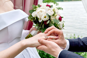 Groom putting wedding ring on bride's finger at wedding ceremony outdoors, close up. Wedding rings. Bouquet of flowers in hand