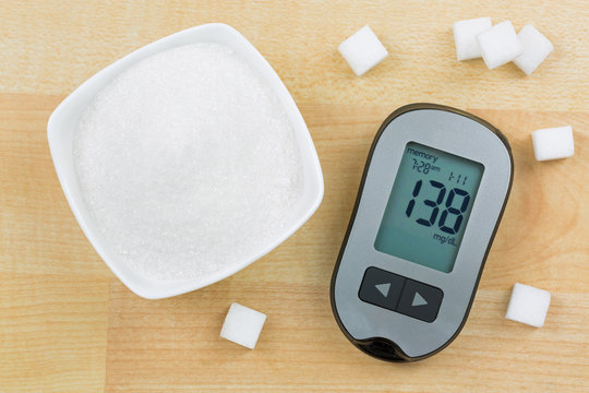 Blood Glucose meter device showing high blood sugar levels, next to bowl of sugar and cubes on wooden background