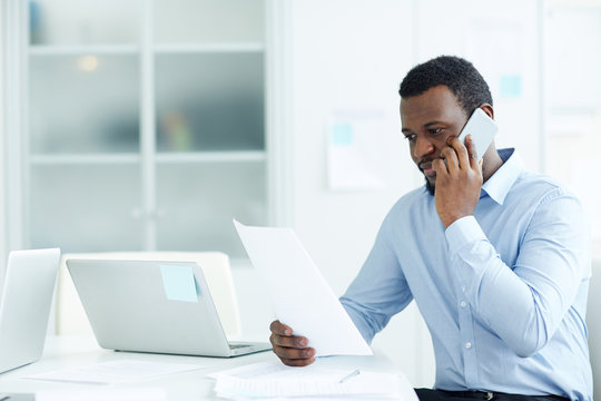 Young African-american Male In Formal Wear Sitting At Desk With Laptops Talking On Phone Looking At Paper In Office