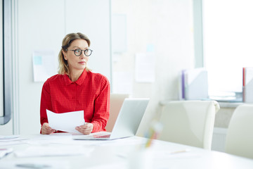 Beautiful emotionless woman in red shirt and eyeglasses sitting at table with papers in white light office looking away. 