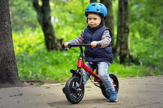 A boy in sleeveless jacket and blue helmet riding runbike in a park