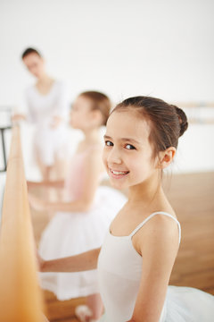 Little ballerina in white clothes staying at wooden machine in studio with people on background
