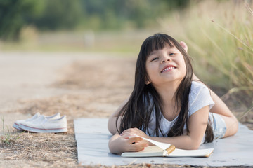 A little girl is lying on her stomach reading on the grass. She has a look of enjoyment on her face and she looks very relaxed. There is quite a bit of negative space around her.