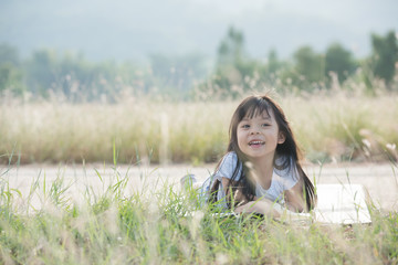 A little girl is lying on her stomach reading on the grass. She has a look of enjoyment on her face and she looks very relaxed. There is quite a bit of negative space around her.