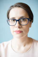 Attractive adult woman in stylish glasses looking at camera while standing on light blue background. 