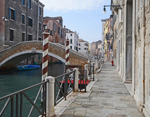 Venice, Italy, medieval stone street along a canal with ancient buildings, a bridge and the typical white and red poles of the gondola docking