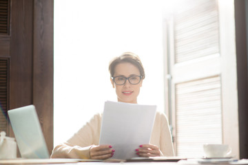 Elegant woman in glasses sitting at cafe table and looking through important documents.