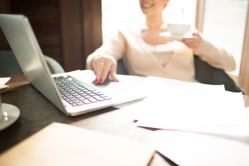 Crop woman with cup of hot beverage sitting at cafe table and browsing laptop. 