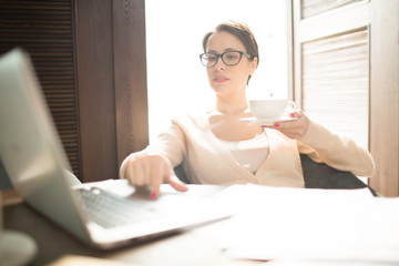 Attractive businesswoman holding cup of aromatic hot beverage and browsing laptop while sitting at cafe table. 