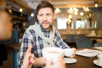 Handsome man in casual outfit drinking hot beverage and listening to colleague while sitting at cafe table together.