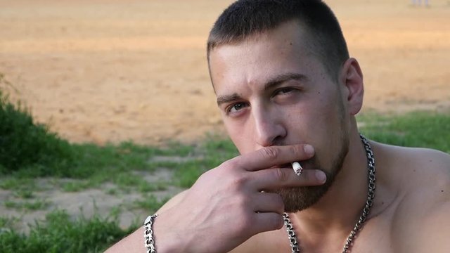 Young attractive strong body guy portrait smoking cigarette exhaling smoke in a park