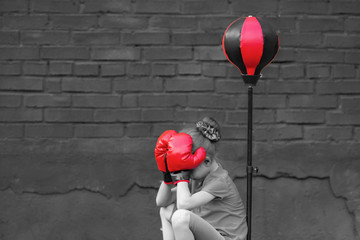A little girl is sitting near the punching bag covering her face with boxing gloves. Black and...