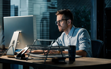 Young focused man doing overtime job watching computer at desktop in dark office. 