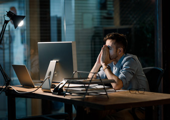 Man in shirt leaning on hands with eyes closed sitting at working table in dark office working...