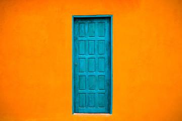 Vivid bright orange colour facade with blue-green closed door in the center of large empty orange wall texture background space.