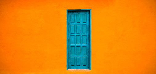 Vivid bright orange colour facade with blue-green closed door in the center of large empty orange wide wall texture background space in panorama format.