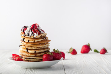 Stack of pancakes with strawberries, whip cream and chocolate syrup on a white plate on a white background. Copy space.