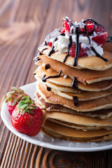  Stack of pancakes with strawberries, whip cream and chocolate syrup on a white plate on a wooden background. Copy space.