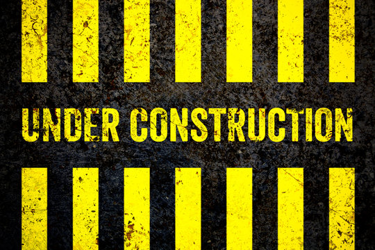 Under construction warning sign with yellow and black stripes painted over cracked concrete wall coarse texture background. Concept for do not enter the area, caution, danger, construction site.