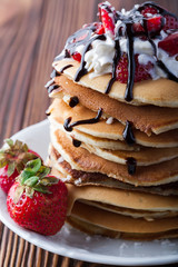  Stack of pancakes with strawberries, whip cream and chocolate syrup on a white plate on a wooden background. Copy space.