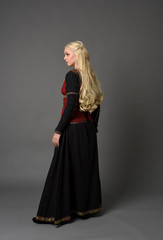 Fototapeta premium full length portrait of pretty blonde lady wearing a red and black fantasy medieval gown. standing pose on grey background.