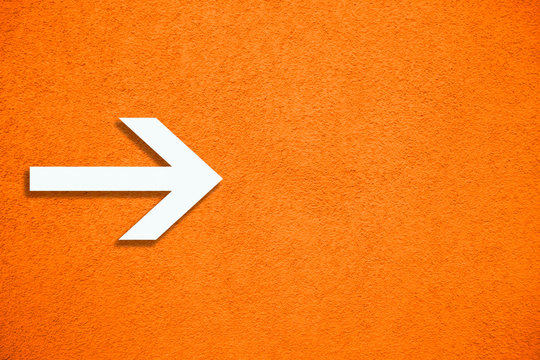White arrow direction sign over vivid bright orange color stucco rough wall as an empty rustic and simple background texture with empty space.