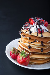  Stack of pancakes with strawberries, whip cream and chocolate syrup on a white plate on a black background. Copy space.
