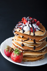  Stack of pancakes with strawberries, whip cream and chocolate syrup on a white plate on a black background. Copy space.