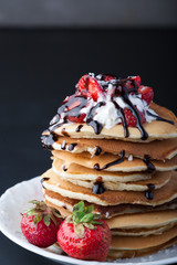 Stack of pancakes with strawberries, whip cream and chocolate syrup on a white plate on a black background. Copy space.
