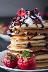Stack of pancakes with strawberries, whip cream and chocolate syrup on a white plate on a wooden background. Copy space.