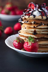 Stack of pancakes with strawberries, whip cream and chocolate syrup on a white plate on a wooden background. Copy space.