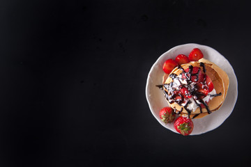 Homemade pancakes with strawberries, whipped cream and chocolate topping, decorated with flowers on black background. Overhead shot. Flat lay. Copy space. Vertical image