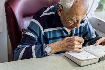 Elderly man reading writings in a notebook with magnifying glass near the window at home